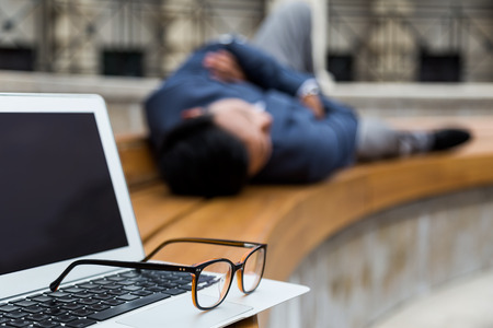 Close up of laptop and glasses. Asian businessman is resting on bench beside them. Concept of much needed rest.の写真素材