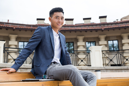 Smiling Asian businessman in dark blue suit is resting on bench near his office during lunch hour. Concept of rest and relaxationの写真素材