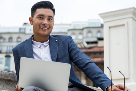 Smiling Asian man in dark blue suit sitting in the street and checking his social media during lunch break in the office. Concept of living in digital worldの写真素材