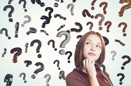 Portrait of young woman in brown sweater standing near question marks floating in the air. Concept...