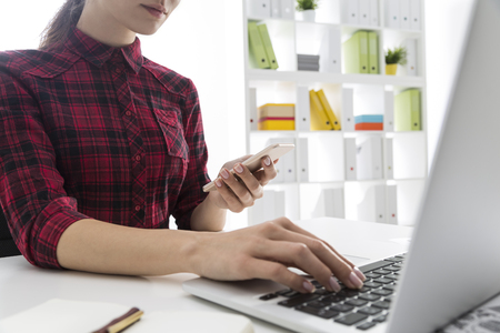 Serious woman in red checkered shirt is working at her laptop keyboard and looking at smartphone. White bookcase with colored folders is seen at the background. Concept of office workの写真素材