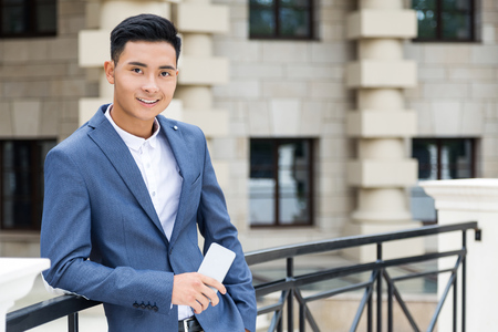 Cheerful Asian businessman standing on the bridge holding his mobile phone in one hand. Concept of confidence and good looks.の写真素材
