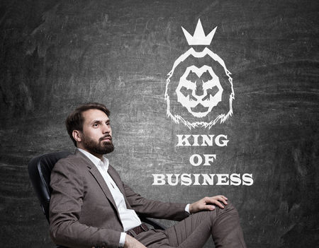 Side view of businessman in brown suit sitting against blackboard with king of business sketch on it.の写真素材