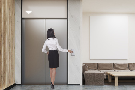 Woman waiting for the elevator in company lobby with vertical poster, coffee table, sofa and elevator. Concept of modern waiting area design. 3d rendering. Mock up.の写真素材
