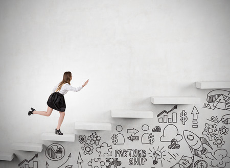 Businesswoman is running up the stairs in concrete wall. Startup icons are drawn with marker under it. Concept of success in business and climbing career ladder. Mock upの写真素材