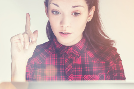 Close up of pretty young girl in red shirt has idea. She is sitting at her laptop and looking at the screen. Concept of insight. Toned image.の写真素材
