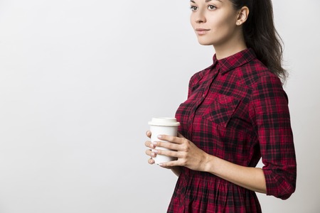 Serious young woman in red checkered dress holding coffee cup with both hands. Concept of being lost in thoughts. Mock upの写真素材