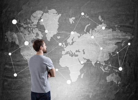 Rear view of businessman in T-shirt looking at world map with placeholders at chalkboard. Concept of international trade.の写真素材