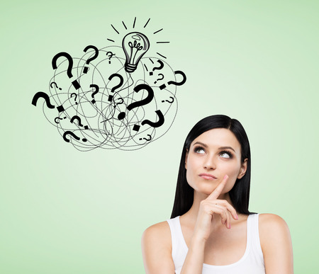 Woman in white tank top is standing near green wall with multiple question marks and light bulb sketch on it. Concept of looking for an answer.の写真素材