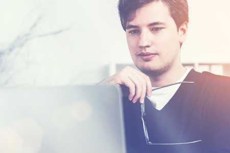 Portrait of thoughtful young businessman with glasses in hand looking at laptop screen. Toned image