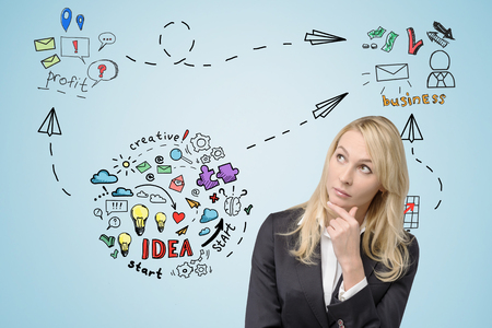 Pensive blond woman is standing near light blue wall with business idea sketch and rubbing her chinの写真素材