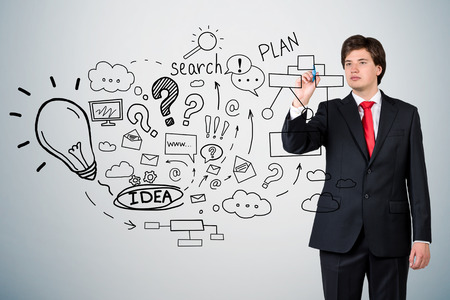 Young businessman wearing red tie is standing near gray wall with startup idea sketch on it. Concept of business planningの写真素材