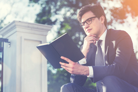 Thoughtful young businessman in suit reading book outside. Concrete columns and trees in the background. Toned imageの写真素材