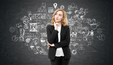 Portrait of a gorgeous businesswoman with red hair standing near the blackboard with business icons on it. Concept of business foundationの写真素材