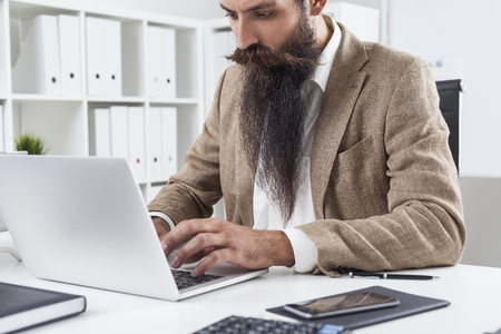 Portrait of businessman with long beard typing at his laptop keyboard in a white office. Concept of office workの写真素材