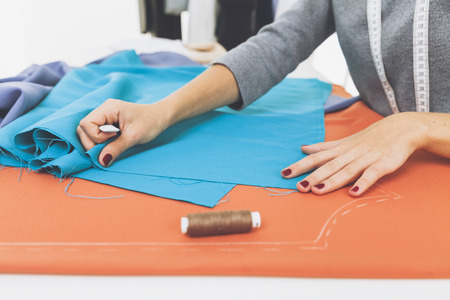 Side veiw of woman's hands drawing a pattern on bright blue material at her workplace in a tailor...
