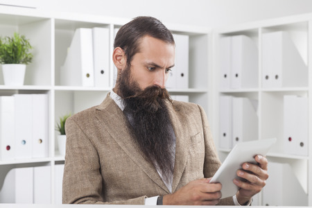 Long bearded businessman is looking at his tablet screen while sitting at his workplace in office with white bookshelves.の写真素材