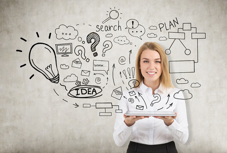 Smiling blond girl is holding a tablet with both hands and looking at the viewer while standing near a concrete wall with internet search sketchesの写真素材