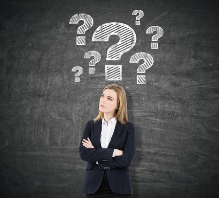 Blond woman is standing with her arms crossed near a blackboard with question mark sketches drawn above her headの写真素材