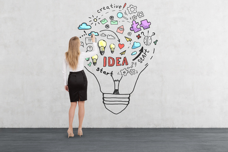 Rear view of blond woman drawing business idea sketch inside a giant light bulb on concrete wall. Concept of creative thinkingの写真素材