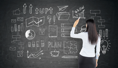 Rear view of a businesswoman with long black hair who is drawing a business idea sketch on a blackboardの写真素材