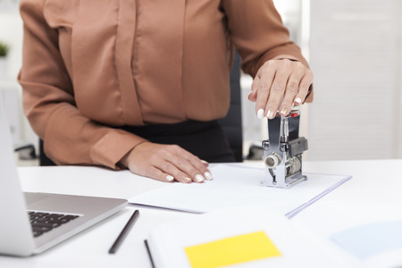 Woman in brown blouse using a stamp on a document. She is sitting in a white office with her laptop...