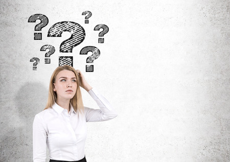 Blond woman in shirt is scratching her head while standing near a concrete wall with question marks....