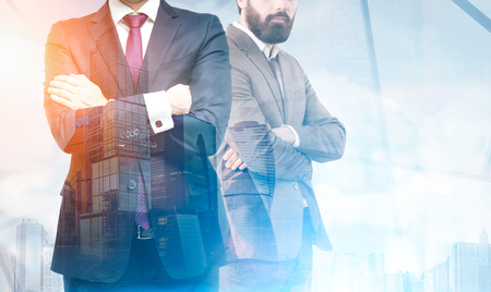 Two business partners are standing together in an office. Their arms are crossed. One is bearded. Cityscape. Toned image. Mock up. Double exposureの写真素材