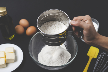 Close up of a hand sewing the flour before making a dough. There are eggs, butter and milk on the...
