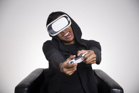 African American guy in a black hoodie is playing a virtual reality game. His tongue is stuck out. He is holding a controller and sitting in a leather armchair. Mock upの写真素材