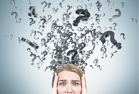 Close up of a blond panicking woman near a gray wall with question marks flying out of it. Concept of challenging issuesの写真素材