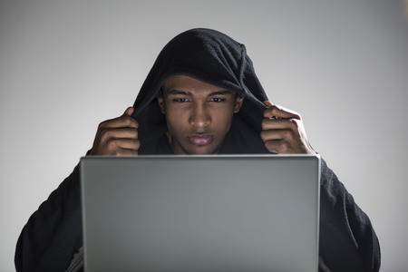 Close up of an African American hacker in a hoodie putting his hood on and looking at his laptop screen. Concept of coding. Mock upの写真素材