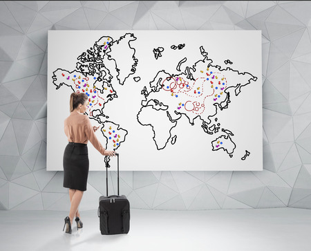 Rear view of a blond businesswoman with a black suitcase standing and looking at a world map with placemarks on it. Concept of travelling.の写真素材