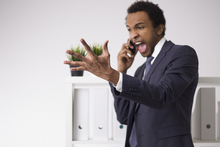 Portait of a frustrated African American businessman yelling while on his phone and gestering franticly. Concept of anger issues and incompetenceの写真素材