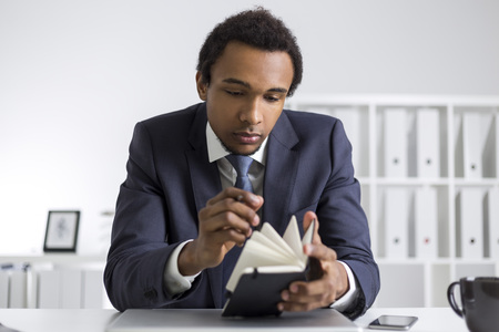 Portrait of a busy young African American gentleman. He is in his office and looking through his notebook. Concept of schedule and time managementの写真素材