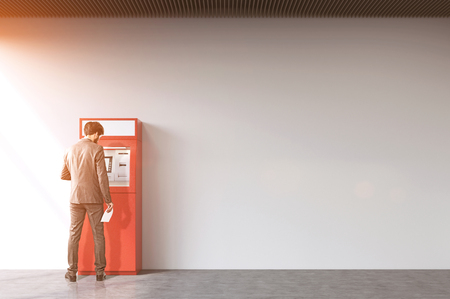 Rear view of a businessman withdrawing cash from a red ATM machine. Concept of financial operations. Mock up. Toned imageの写真素材