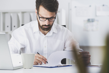 Portrait of a young man in a white shirt and glasses who is signing a document in his office. There is a laptop and a coffee cup on his desk.の写真素材