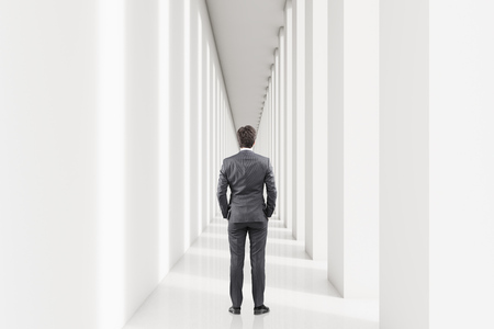 Rear view of a young businessman in a suit is standing in a white corridor and thinking. Concept of management. 3d rendering. Mock upの写真素材