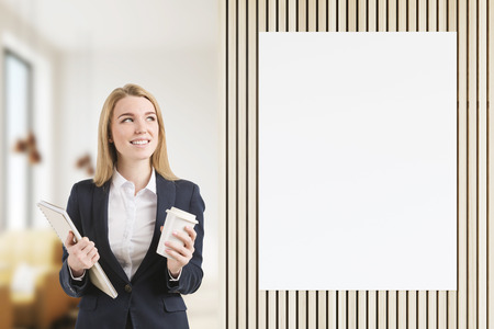 Beautiful blond woman with a cup of coffee and a notebook standing in a restaurant near a blank poster. 3d rendering. Mock up. Toned imageの写真素材