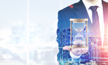 Close up of a businessman holding hourglass. Office. Night cityscape. Concept of time management. Mock up. Toned image. Double exposureの写真素材