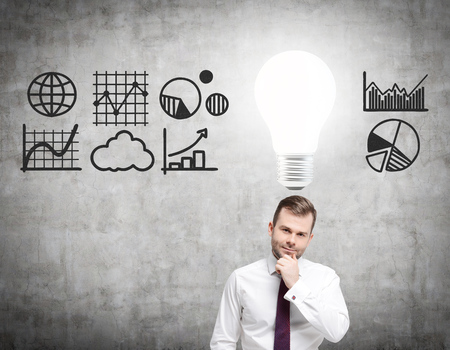 Thoughtful bearded businessman is standing near a concrete wall with graphs drawn on it. There is a large glowing light bulb above his head. Concept of decision making.の写真素材