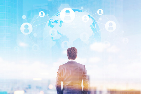 Rear view of a businessman in a suit looking at a network sketch against a globe in a sky. Cityscape is in the background. Double exposure. Toned image.の写真素材