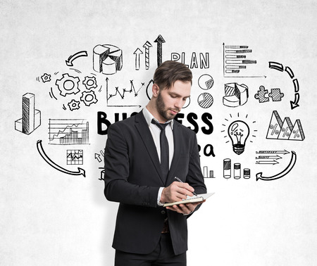 Close up of a bearded businessman with a notebook writing while standing near a concrete wall with business idea iconsの写真素材