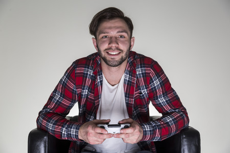 Portrait of a cheerful young man in a checkered shirt sitting in a leather armchair and holding a video game controller.の写真素材