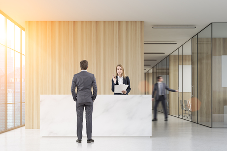 Man standing near a reception desk talking to a receptionist. His colleague is closing a meeting room door. 3d rendering. Mock up. Toned image.の写真素材