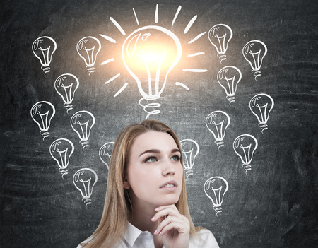 Close up of a pensive blond womans head near a blackboard with one big and shining light bulb above her and many smaller ones.の写真素材