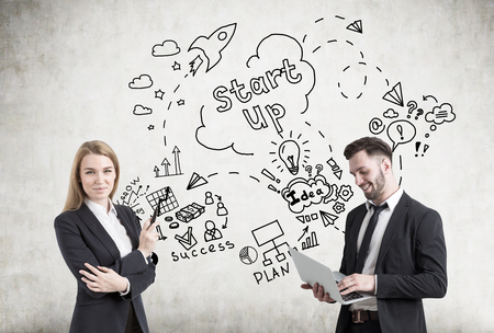 Portrait of a businesswoman with a marker and a businessman with a laptop standing near a concrete wall with a start up sketch.の写真素材