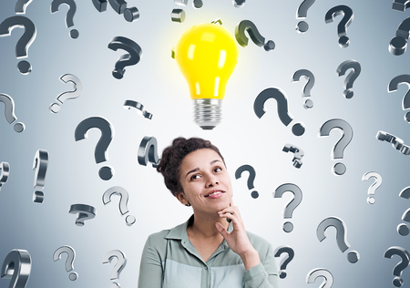Portrait of an African businesswoman standing near a gray wall with a bright light bulb icon and question marks falling behind her.の写真素材