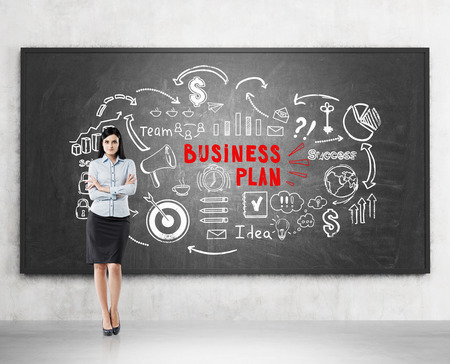 Portrait of a black haired woman standing with crossed arms near a blackboard with a white and red business plan sketch on it.の写真素材