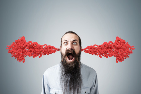 Portrait of an angry bearded man standing near a gray wall. There are two arrows made of question marks coming out of his ears.の写真素材
